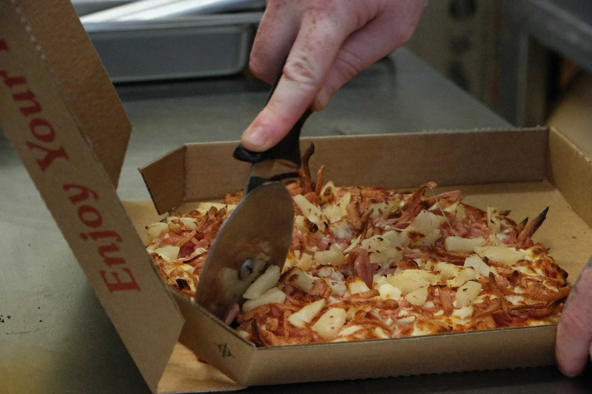 Hands slicing ham and pineapple pizza with wheel cutter in takeaway box